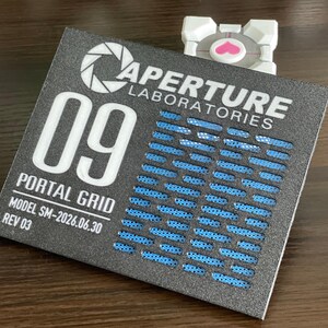May include: A rectangular black sign with white text reading APERTURE LABORATORIES, 09, PORTAL GRID, MODEL SM-2026.06.30, and REV 03. The sign features a blue grid pattern and a white circular logo. A small white cube with a pink heart sits behind it.