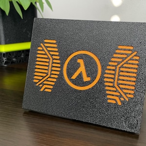 Puede incluir: Una placa de exhibición rectangular negra con superficie texturizada que presenta un símbolo Lambda naranja central flanqueado por dos patrones naranjas similares a circuitos. El diseño está iluminado desde atrás, creando un contraste.