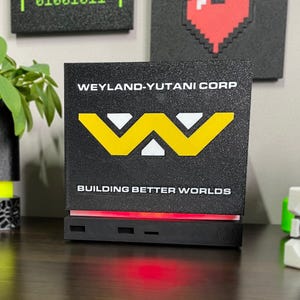 May include: A black rectangular display sign featuring the white text WEYLAND-YUTANI CORP and BUILDING BETTER WORLDS with a large yellow and white W logo in the center. The base of the sign emits a bright red light glow onto the surface below.