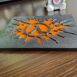 Puede incluir: Un posavasos cuadrado con una capa superior negra que presenta un recorte del símbolo lambda de Half-Life y líneas dentadas radiantes, revelando una capa base texturizada de color naranja brillante debajo del diseño.