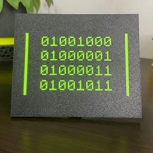 Front view of the black plaque with green binary code spelling HACK.