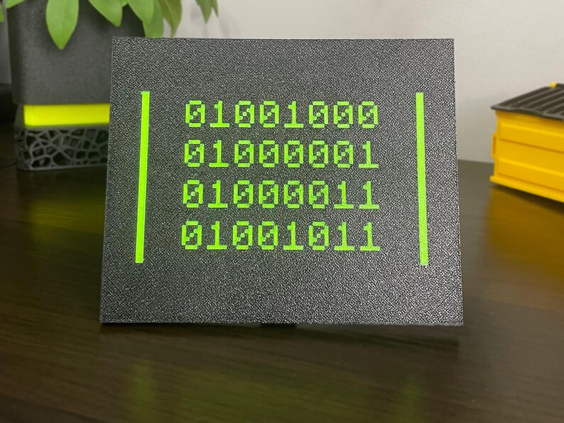 Front view of the black plaque with green binary code spelling HACK.