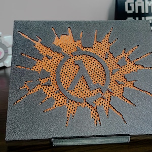 May include: A rectangular black plaque featuring a cutout of the Half-Life lambda symbol centered within a jagged, starburst-shaped opening. The cutout reveals a bright orange, textured mesh background behind the dark, grainy surface.