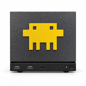 Può includere: Un dispositivo elettronico rettangolare nero dalla superficie ruvida, caratterizzato da un personaggio giallo pixelato simile a un alieno sul pannello frontale. La base include due porte USB, una fessura orizzontale, una spia bianca e un tasto.