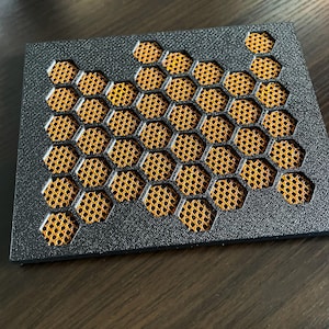 May include: A rectangular black coaster featuring a textured surface with a cut-out honeycomb pattern. Beneath the black frame, a fine orange mesh grid is visible through the hexagonal openings, creating a layered contrast in color and texture.