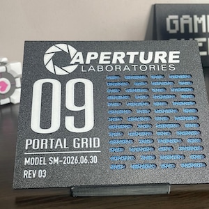 May include: A black rectangular sign featuring the Aperture Laboratories logo, the number 09, and the text Portal Grid, Model SM-2026.06.30, Rev 03. The right side has a grid pattern revealing a blue textured layer behind the black surface.