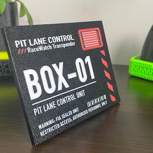 May include: A rectangular black sign with white text reading PIT LANE CONTROL RaceWatch Transponder, BOX-01, and PIT LANE CONTROL UNIT. It features a red square graphic with horizontal lines, red diagonal stripes, and a barcode graphic at the bottom.