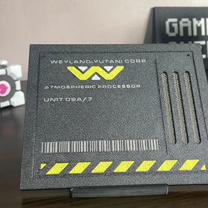 Könnte beinhalten: Eine rechteckige schwarze Plakette mit dem Text WEYLAND-YUTANI CORP, ATMOSPHERIC PROCESSOR und UNIT 09A/7. Das Design umfasst ein gelbes Logo, drei vertikale Lüftungsschlitze, einen Strichcode und gelbe diagonale Warnstreifen am unteren Rand.