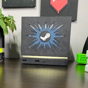 May include: A black rectangular electronic device stand featuring a central cutout with a blue mesh background and a white Steam logo. The stand has a textured black front panel, a gold-coloured horizontal accent strip, and a base with three ports.