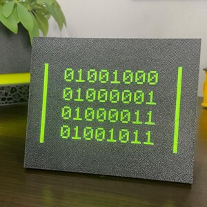 HACK binary plaque displayed upright beside a plant on a gaming desk.