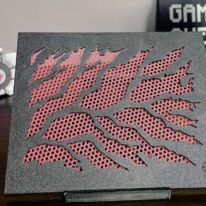 May include: A rectangular black panel featuring a jagged, claw-mark style cutout pattern that reveals a pink mesh layer underneath. The surface has a textured finish, and a small circular logo is embossed in the bottom right corner of the panel.