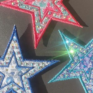 May include: Three star-shaped fabric patches featuring floral patterns in pink, blue, and light blue. Each patch is outlined with thick embroidery thread and embellished with small, reflective rhinestones scattered across the surface.