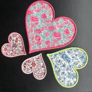 May include: Four heart-shaped fabric patches with floral patterns. The largest heart has a pink border. The other hearts have similar designs with different colours and sizes. One heart has a blue and white floral pattern.