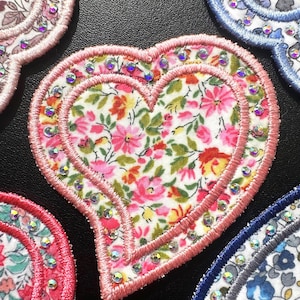 May include: A heart-shaped fabric patch featuring a white background with a colourful floral pattern of pink, red, and green flowers. The patch is bordered by a thick pink embroidered edge and accented with small, clear, reflective rhinestones.