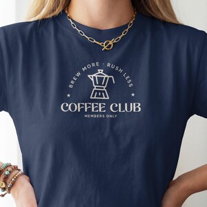 Könnte beinhalten: Ein marineblaues T-Shirt mit einer weißen Grafik einer Mokkakanne. Der Text "BREW MORE RUSH LESS" steht in einem Bogen über der Kanne, während "COFFEE CLUB" und "MEMBERS ONLY" in weißer Schrift unter dem Kaffeesymbol gedruckt sind.