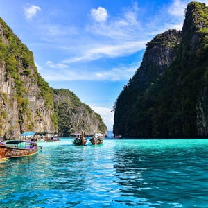 May include: A scenic view of a tropical bay with turquoise water and several longtail boats. Towering cliffs covered in lush green vegetation frame the bay under a partly cloudy blue sky. The boats are brown and colorful.