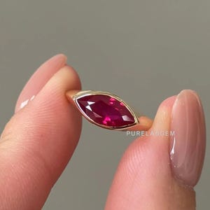 Op de afbeelding: Een gouden ring met een marquise-geslepen robijn. De robijn heeft een dieprode kleur en is gezet in een bezel setting. De ring wordt tussen twee vingers gehouden tegen een neutrale achtergrond. De tekst "PURELABGEM" is zichtbaar.
