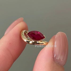 May include: A gold ring featuring a marquise-cut ruby gemstone. The ring has a smooth, polished band and a setting that cradles the vibrant red stone. The text "PURELABGEM" is visible on the ring.
