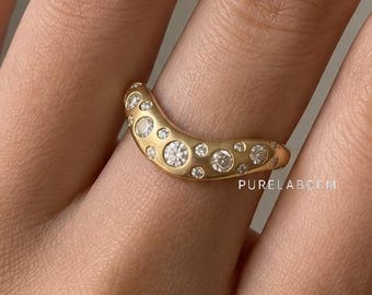 May include: A curved gold band ring featuring a scattered arrangement of multiple round, clear, brilliant-cut stones set flush into the metal surface. The ring has a smooth, matte-textured finish and includes the text PURELABGEM on the band.