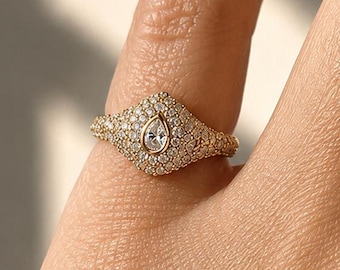 May include: A gold signet ring featuring a central pear-shaped stone surrounded by a pavé setting of numerous small, round, clear stones. The band and the face of the ring are entirely covered in these small, sparkling stones, creating a textured surface.