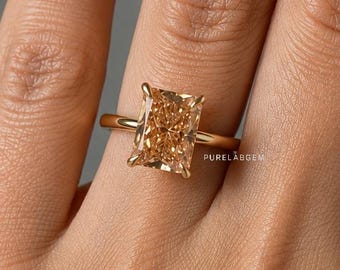 May include: A gold ring featuring a large, rectangular, radiant-cut champagne-colored gemstone set in a four-prong setting. The band is a simple, polished gold metal. The text PURELABGEM is visible in small white letters near the gemstone setting.