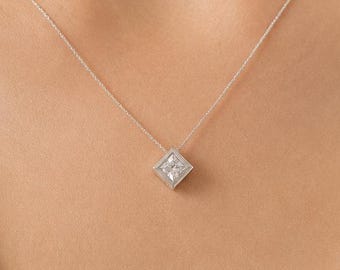 May include: A delicate silver-toned chain necklace featuring a small, square-shaped pendant with a central clear stone. The pendant is set in a metallic frame, suspended from a thin, polished metal chain that rests against the collarbone area.