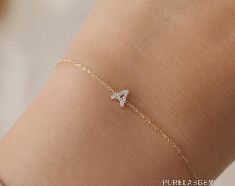 May include: A delicate gold chain bracelet featuring a single, small letter A pendant encrusted with tiny clear stones. The letter A is centered on the thin chain, which is secured around the wrist. The text PURELABGEM is visible on the skin.