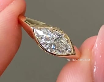 May include: A gold ring featuring a marquise-cut diamond. The diamond is set in a bezel setting, and the band is a simple, polished gold. The ring is held up against a neutral background.