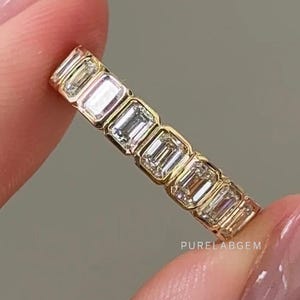 May include: A gold band ring featuring a series of rectangular, clear gemstones. The ring is held between two fingers against a neutral background. The text "PURELABGEM" is visible on the ring.