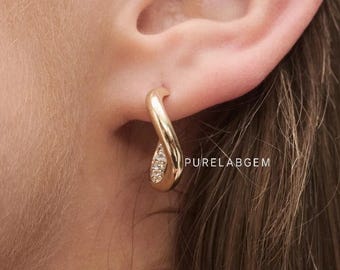 May include: A gold hoop earring with a twisted, ribbon-like design featuring a small section of embedded clear stones. The earring is worn in the earlobe and displays the text PURELABGEM in small white letters near the lower curve of the metal.