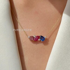 Peut inclure: Un collier en or délicat orné de trois pierres précieuses en forme de goutte, roses, rouges et bleues, rehaussées de petites pierres blanches. Le collier est présenté sur un fond blanc, avec le texte "PURELABGEM" visible.
