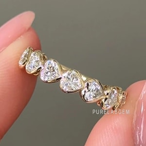 May include: A gold ring featuring a series of heart-shaped diamonds. The ring is held between two fingers against a neutral background. The ring's design is delicate and elegant, showcasing the sparkle of the diamonds.