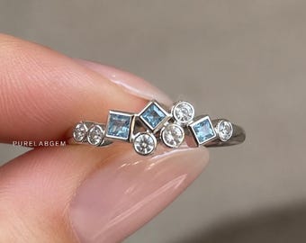 May include: A silver-toned ring featuring a cluster of alternating round and square-cut stones. The square stones are light blue, while the round stones are clear. The text PURELABGEM is printed on the band of this jewelry piece.