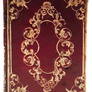 May include: Antique book with a deep red cover and ornate gold filigree designs. The cover features a central oval design and decorative corner elements. The edges of the book are also trimmed in gold.