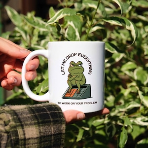 Peut inclure: Un mug en céramique blanc orné d'une illustration de grenouille verte grincheuse assise sur une calculatrice, les bras croisés. Le texte au-dessus de la grenouille indique "LET ME DROP EVERYTHING" et le texte en dessous "TO WORK ON YOUR PROBLEM."