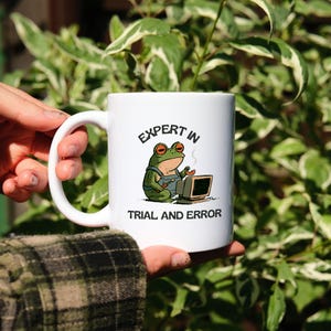 May include: A white ceramic mug featuring a cartoon illustration of a green frog wearing blue overalls, sitting at a computer, and holding a small snack. The text above the frog reads EXPERT IN and below the frog reads TRIAL AND ERROR.