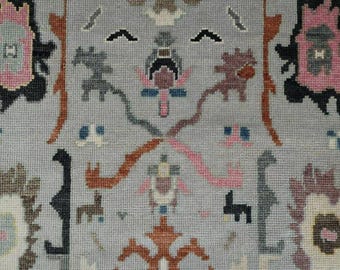 May include: Close-up of a woven textile with a light gray background. The design features repeating patterns in shades of pink, brown, black, and blue. The pattern includes stylized animal and floral motifs, creating a textured appearance.