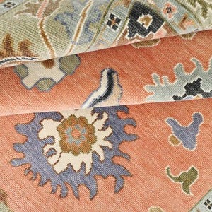 May include: Close-up of a rolled-up area rug with a detailed floral pattern. The rug features a coral background with blue, green, and brown accents. The design includes stylized flowers and geometric shapes, showcasing a traditional style.