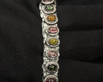 May include: A silver bracelet featuring oval-cut gemstones in various colors, including pink, green, and yellow, each surrounded by a halo of small, clear stones. The bracelet is displayed against a dark background.