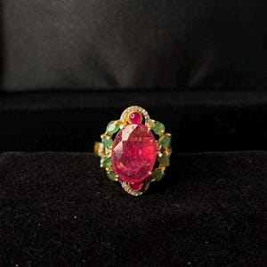May include: A gold ring with a large, oval-cut ruby as the focal point. The ruby is encircled by smaller emeralds and diamonds, with a small ruby at the top. The ring is set against a dark backdrop.
