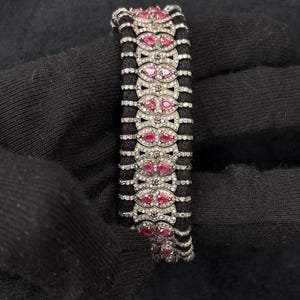 May include: A luxury bracelet featuring alternating rows of black cord and ornate silver links. Each silver link is encrusted with small, clear stones and accented with vibrant, oval-shaped red gemstones. The bracelet is a statement piece of jewelry.