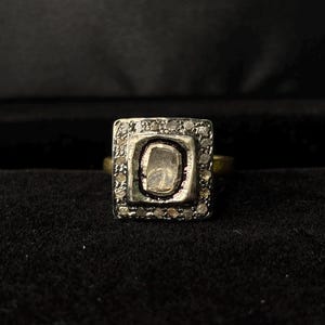 May include: A square ring featuring a central, pale stone. The stone is encircled by smaller, clear stones set in a silver-toned metal. The ring's band is gold-coloured. The ring is displayed against a dark backdrop.