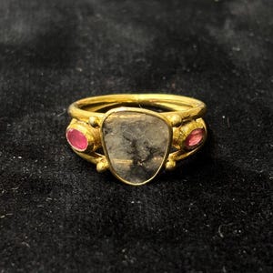 May include: A gold ring with a large, irregularly shaped, gray stone at the center. Two smaller, oval-shaped, red stones are set on either side of the central stone. The ring has a double band.