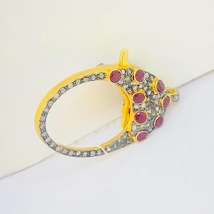 May include: A gold-colored clasp with a rounded oval shape. The clasp is adorned with small, round, red gemstones and tiny, sparkling, gray stones. The clasp is designed to secure jewelry.