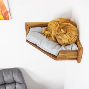 May include: A wooden cat bed mounted on a white wall. The bed has a light gray cushion and a sleeping orange cat. The bed is made of stained wood and is designed to fit in a corner.