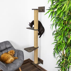 May include: A cat tree with a sisal-wrapped post and three wooden platforms. A black cat is climbing the tree, while an orange cat rests in a gray armchair. The cat tree is mounted on a white wall.