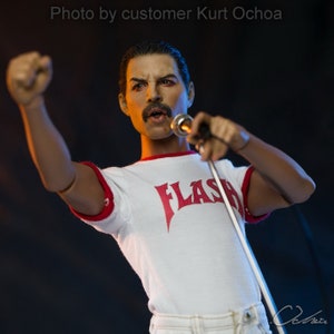 May include: A white t-shirt with red trim and the word "FLASH" printed in red on the front. The shirt is being worn by a person with a moustache and a microphone in their hand.