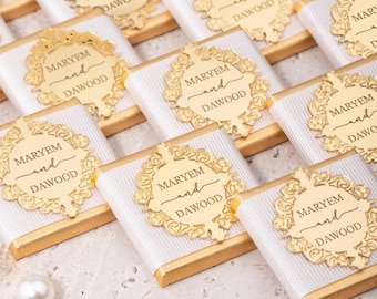 May include: Arrangement of gold-wrapped chocolate bars with white ribbon and gold embellishments. Each bar features a gold decorative element with the names "MARYEM and DAWOOD" in elegant script. A pearl sits in the lower left corner.