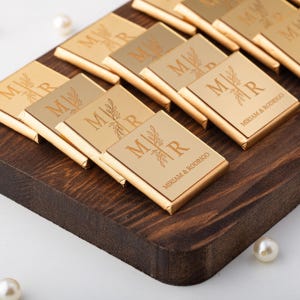 May include: Gold-wrapped chocolate bars on a dark wooden tray. Each bar has the initials "M R" with a leaf design and the names "MIRIAM & RODRIGO". The chocolates are presented with a pearl accent.