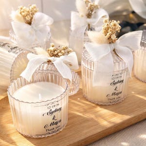 以下が含まれることがあります： 白ワックスキャンドルが入った透明なリブガラスのキャンドルホルダー。各キャンドルは白いリボン、ドライフラワー、テキスト「12/19/26 Sydney & Mason Thank you」のラベルで飾られています。